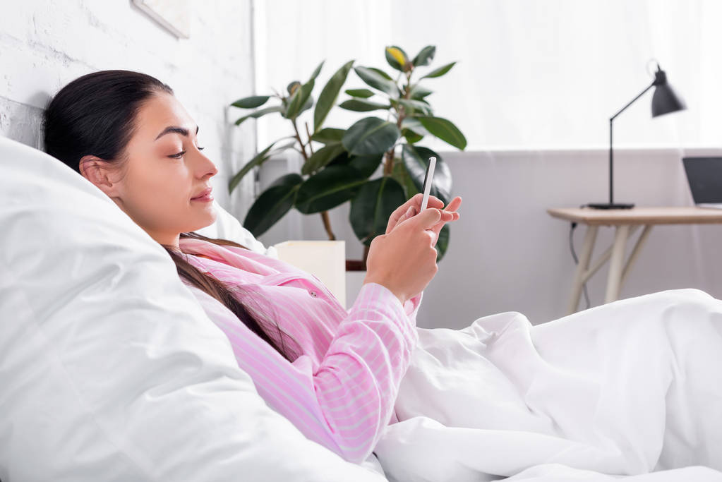 Seitenansicht der attraktiven Frau im Pyjama mit Smartphone, während sie zu Hause im Bett liegt - Foto, Bild