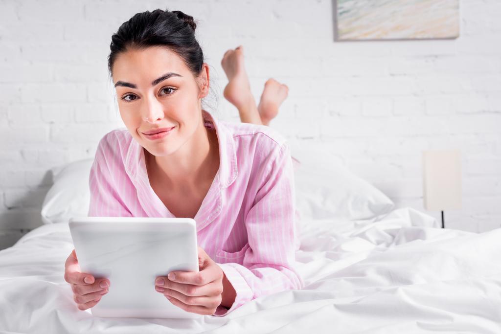 портрет улыбающейся женщины в пижаме с планшетом, лежащей на кровати дома
 - Фото, изображение