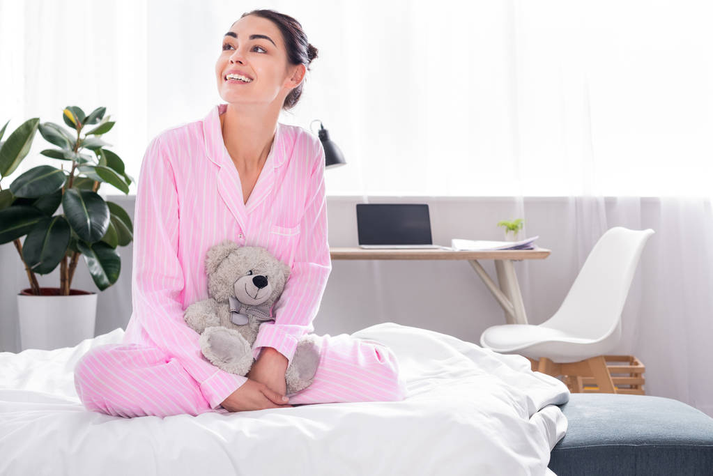 весела жінка в рожевій піжамі з плюшевим ведмедем сидить на ліжку вдома
 - Фото, зображення