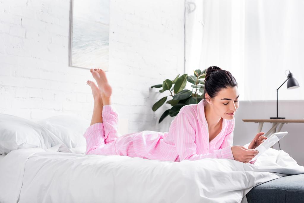 Widok z boku uśmiechający się kobiety w piżamie przy użyciu tabletu leżąc na łóżku w domu - Zdjęcie, obraz
