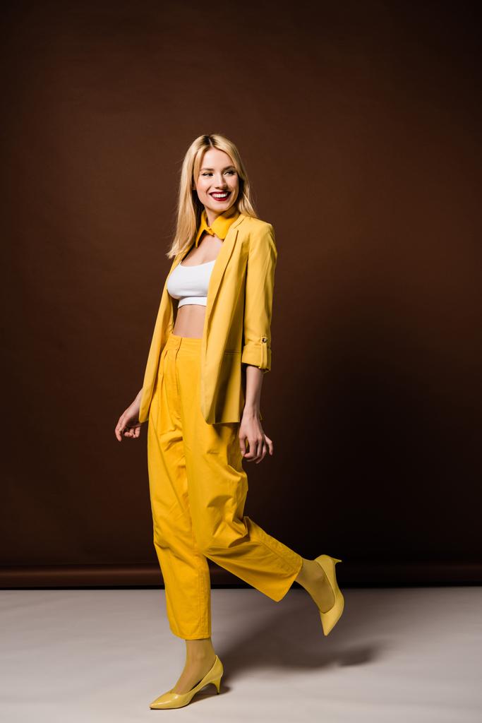full length view of cheerful blonde girl in stylish yellow outfit smiling and looking away on brown - Photo, Image
