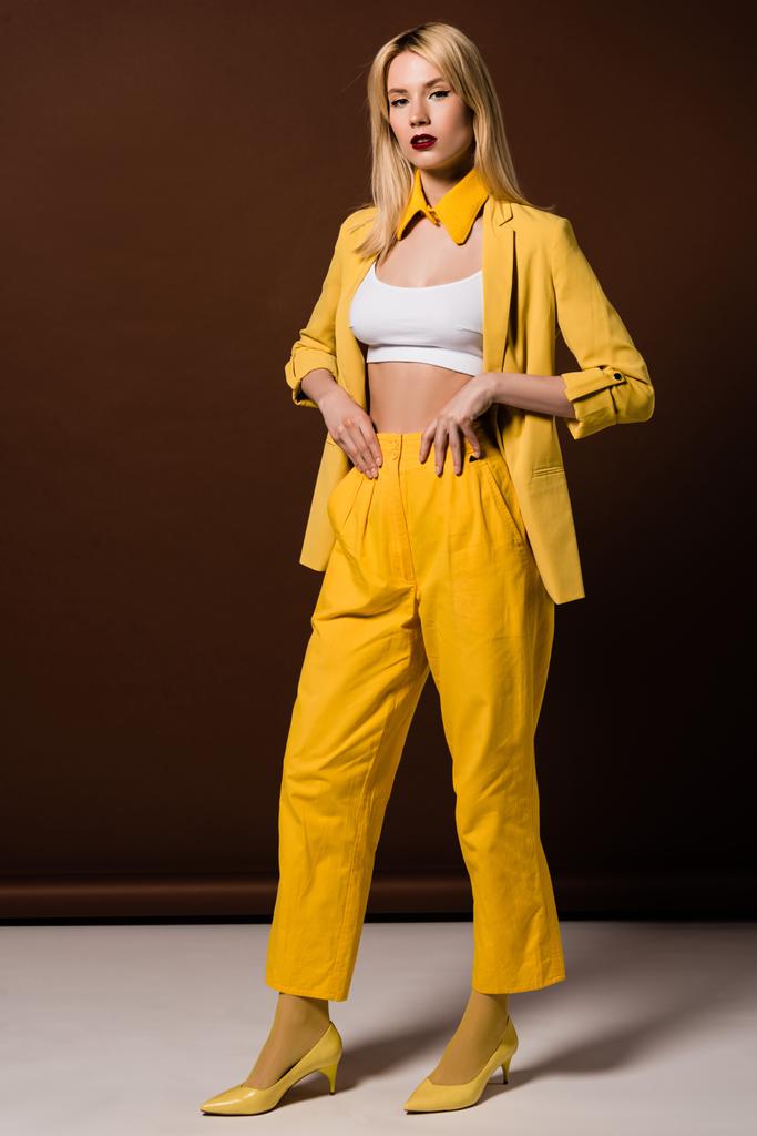 Ganzkörperansicht der schönen blonden Frau in stilvoller gelber Kleidung, die in die Kamera auf braun schaut  - Foto, Bild