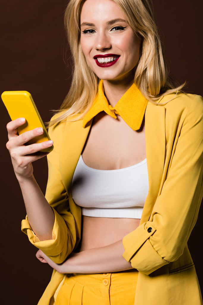 Όμορφη μόδας ξανθιά γυναίκα κρατώντας κίτρινο smartphone και χαμογελαστός στην κάμερα που απομονώνονται σε καφέ - Φωτογραφία, εικόνα