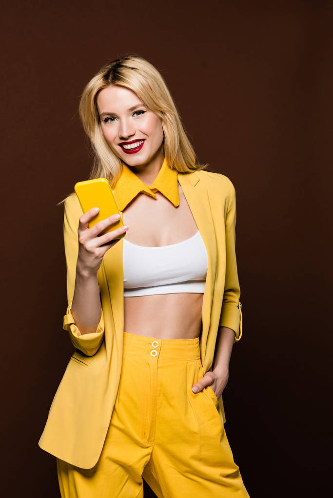 hermosa mujer rubia elegante sosteniendo teléfono inteligente amarillo y sonriendo a la cámara aislada en marrón
 - Foto, Imagen