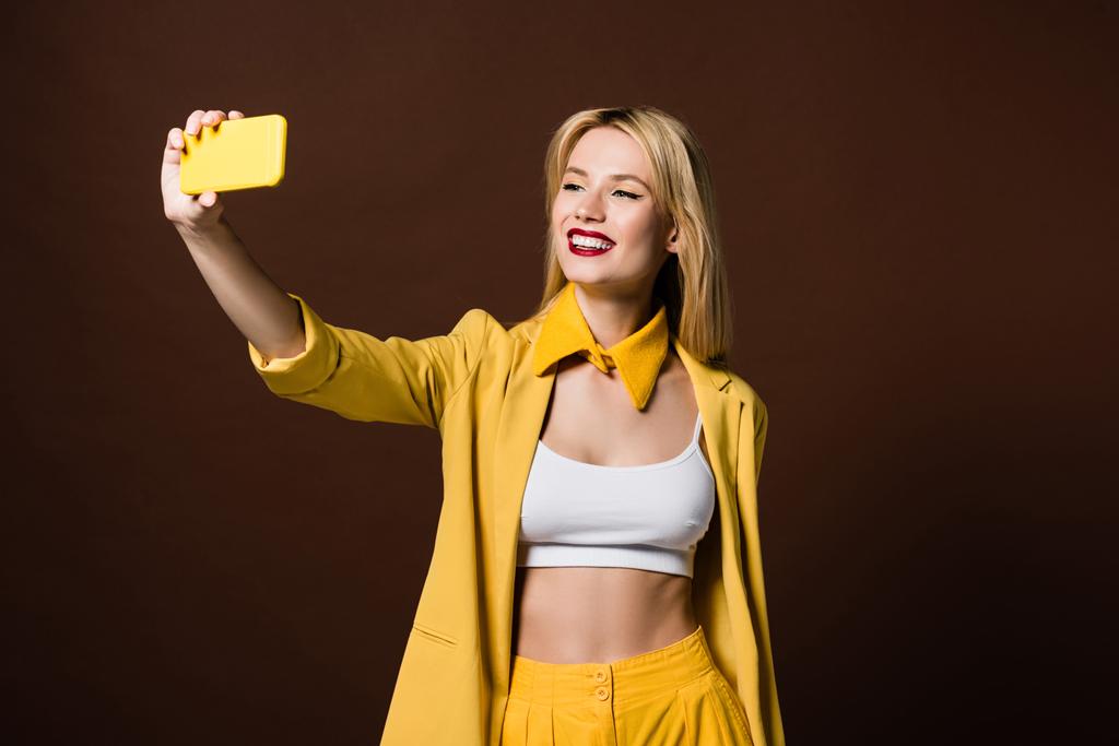 όμορφη χαμογελαστή κομψό ξανθιά κοπέλα λαμβάνοντας αυτοπορτρέτα με κίτρινο smartphone που απομονώνονται σε καφέ - Φωτογραφία, εικόνα