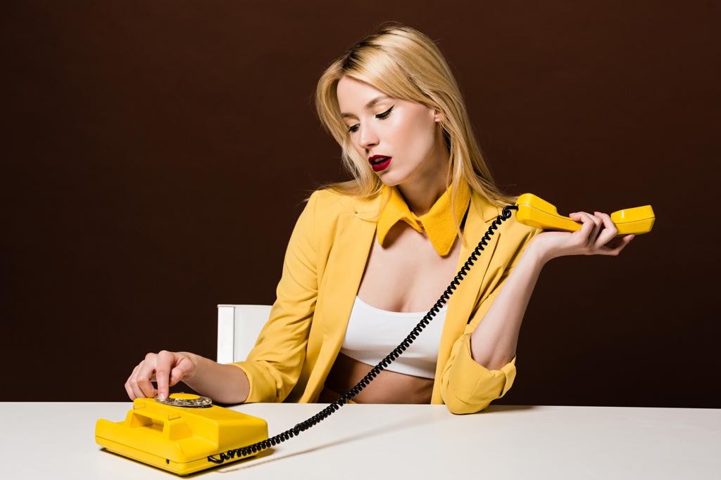 beautiful stylish blonde woman in yellow clothes using rotary phone while sitting on brown - Photo, Image