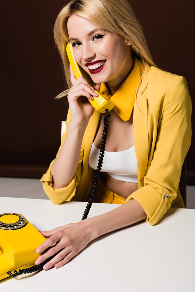 красивая блондинка говорит по желтому винтажному телефону и улыбается в камеру на коричневый
 - Фото, изображение