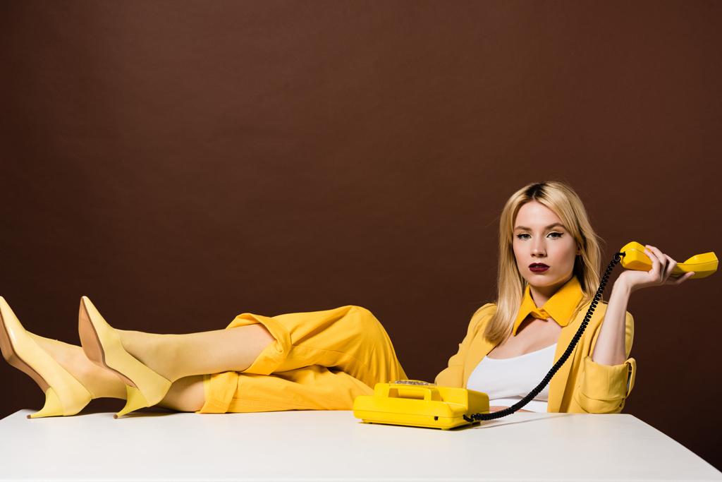 krásná stylová blond dívka drží žlutý sluchátko a při pohledu na fotoaparát na hnědé - Fotografie, Obrázek