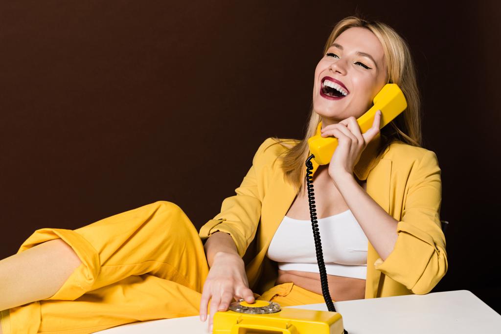 alegre rubia chica hablando por amarillo vintage teléfono y riendo en marrón
 - Foto, imagen