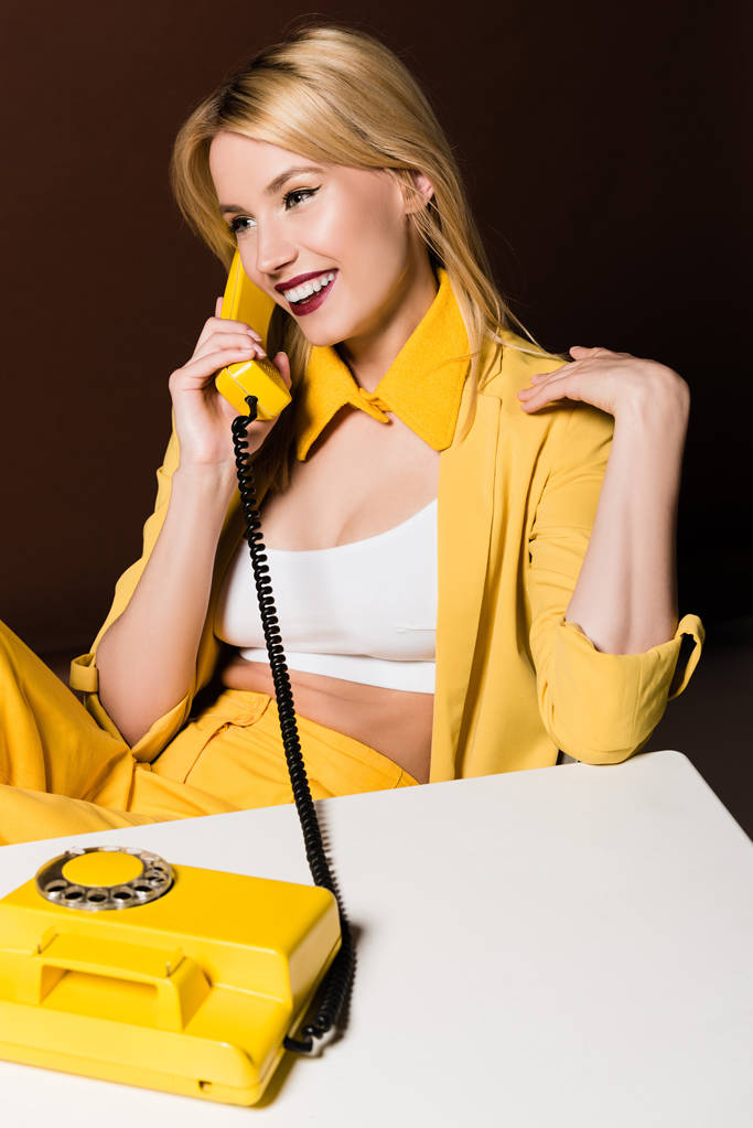 schöne lächelnde stilvolle blonde Mädchen im Gespräch mit gelben Vintage-Telefon auf braun - Foto, Bild