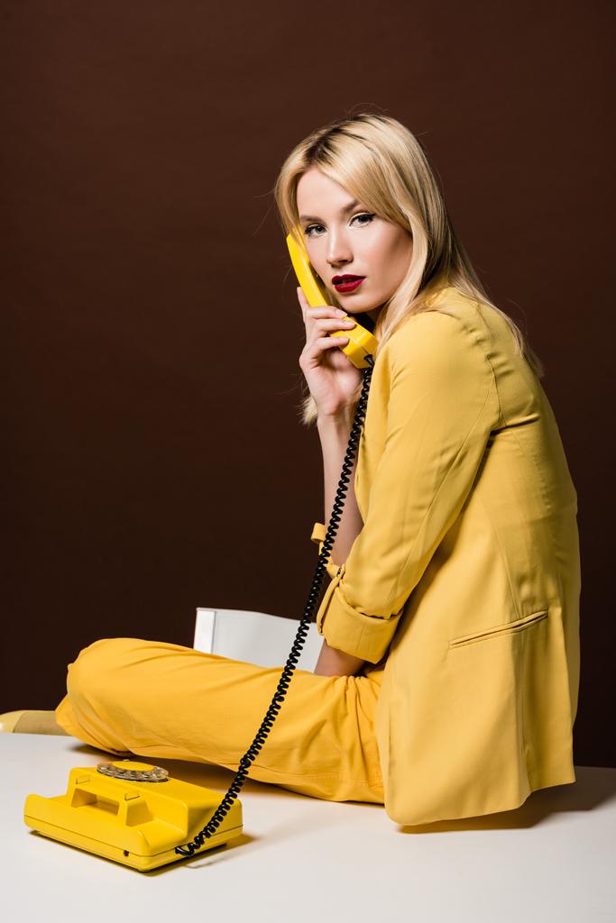 mooie stijlvolle blonde vrouw in gele kleding praten door roterende telefoon en kijken naar camera op brown - Foto, afbeelding