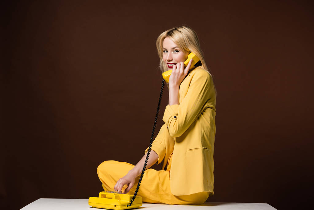 красивая молодая женщина разговаривает по желтому винтажному телефону и улыбается камере на коричневом
 - Фото, изображение