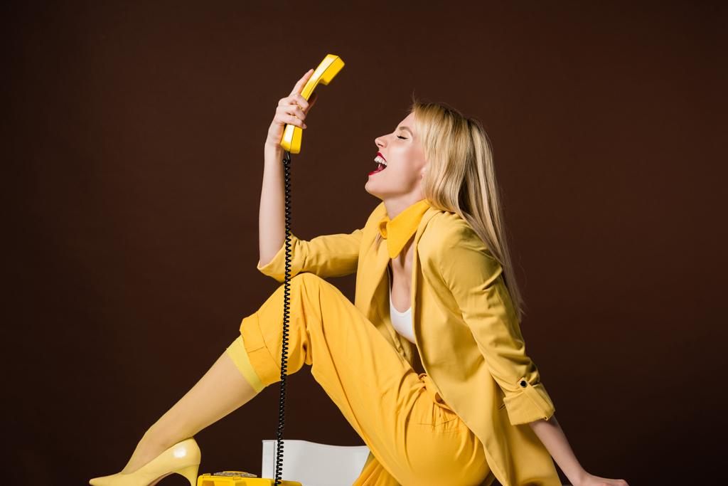 excitada chica rubia elegante sosteniendo el auricular amarillo y sentado en marrón
 - Foto, imagen