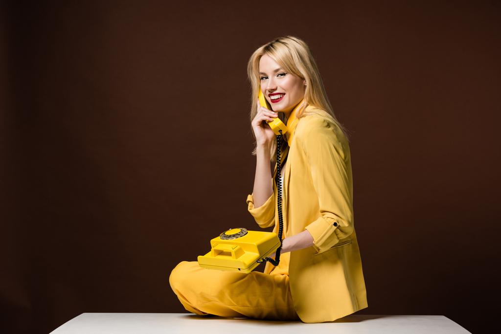 krásná blondýnka v elegantní žluté oblečení drží vintage telefon a usmívá se na kameru na hnědé - Fotografie, Obrázek