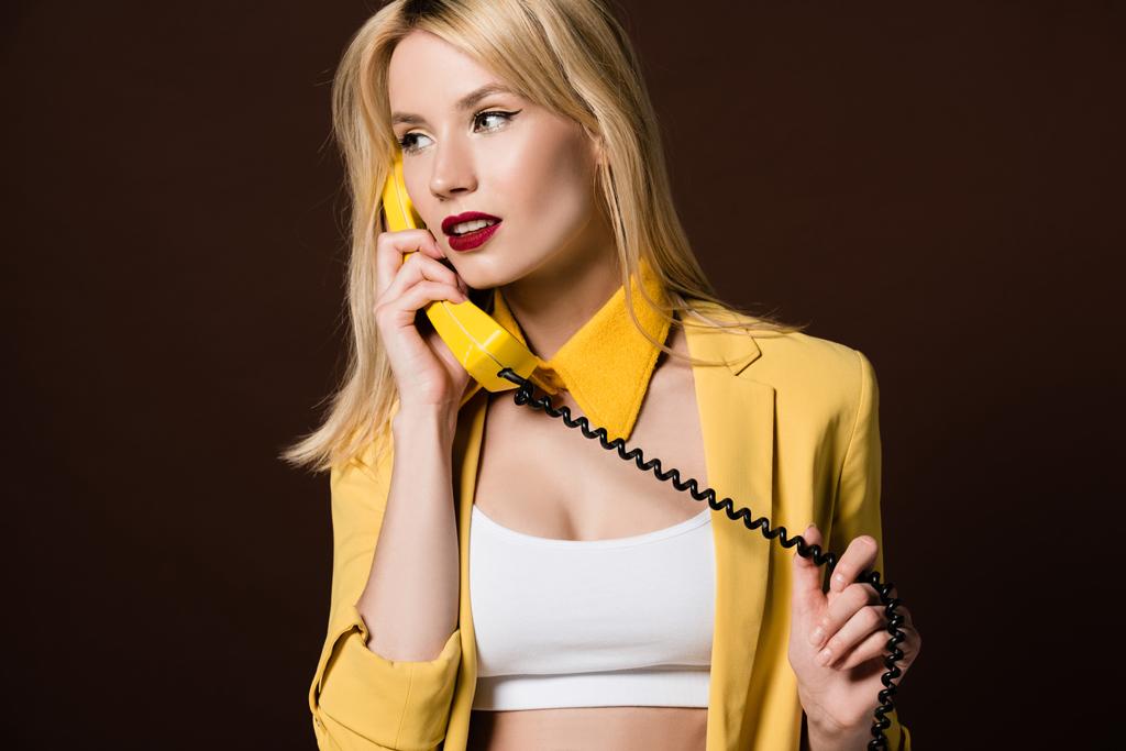 krásná stylová holka mluví o žluté Retro telefon a hledět pryč izolované na hnědé - Fotografie, Obrázek