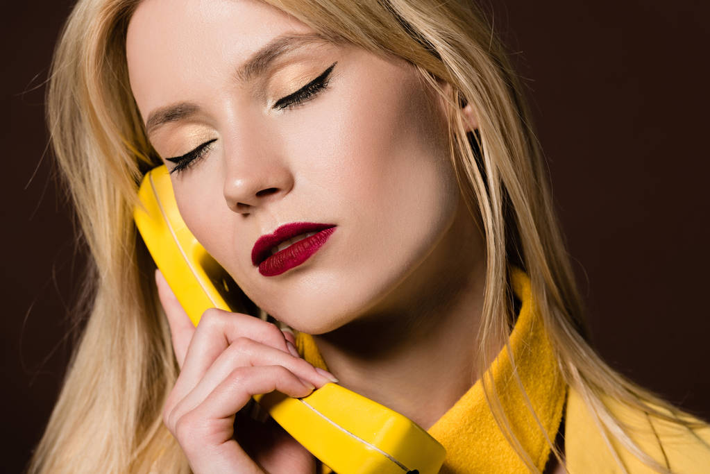 Крупный план красивой стильной блондинки с закрытыми глазами, держащей телефон на коричневом
 - Фото, изображение