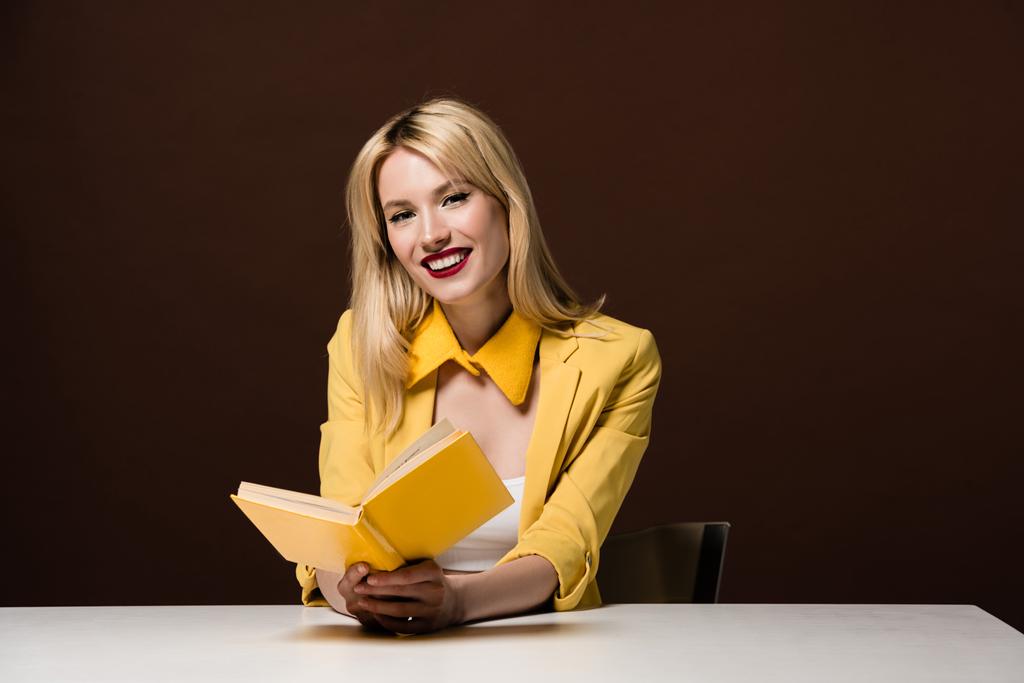 krásná stylová blond dívka žlutý knihu a usmívá se na kameru na hnědé - Fotografie, Obrázek