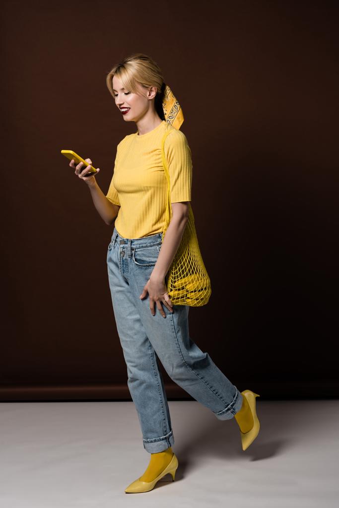 full length view of stylish young blonde woman with bananas in string bag using yellow smartphone on brown - Photo, Image