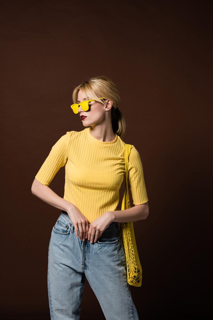 élégante fille blonde avec sac à ficelle portant des lunettes de soleil jaunes et regardant loin isolé sur brun
  - Photo, image