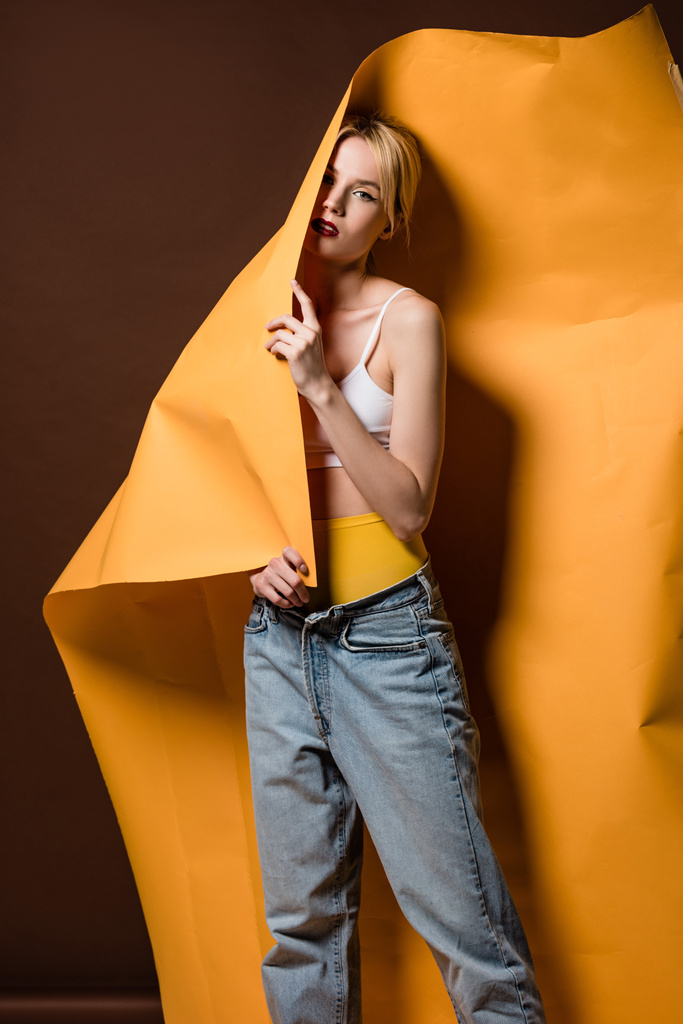 beautiful stylish blonde girl posing with orange paper and looking at camera on brown   - Photo, Image