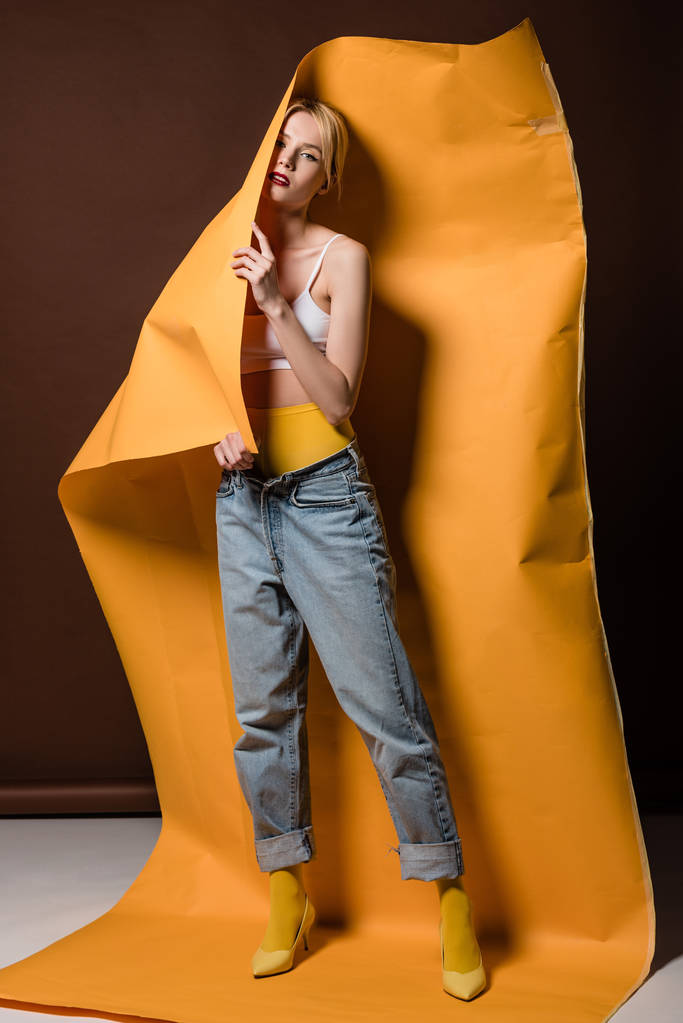 full length view of beautiful stylish blonde girl posing with orange paper and looking at camera on brown  - Photo, Image