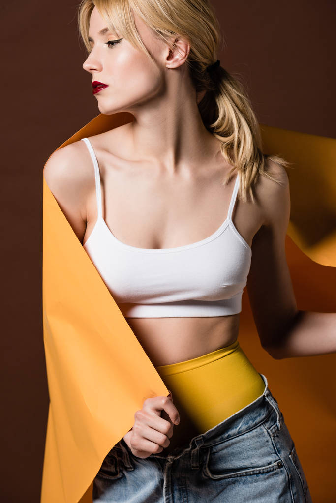 krásná stylová blond dívka zabalená do oranžový papír při pohledu od izolovaných na hnědé - Fotografie, Obrázek