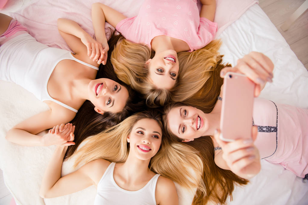 Portrait of sexy, pretty, sweet, charming, funny, foolish, lovely,  cheerful girls lying head to head on bed using smart phone shooting selfie on front camera, holding hands - Photo, Image
