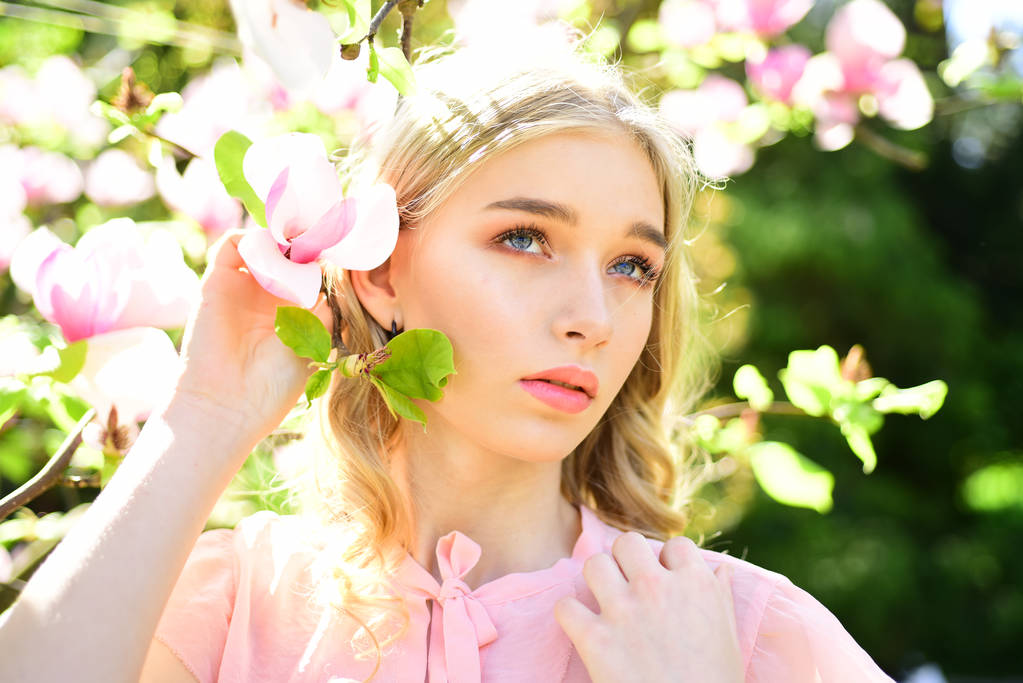 hübsches Mädchen in rosa Bluse posiert neben blühendem Baum. blonde junge Frau mit zartem Zweig und wunderschöner Magnolienblüte. Entspannter Spaziergang an warmen, sonnigen Tagen, Frühlingskonzept. - Foto, Bild