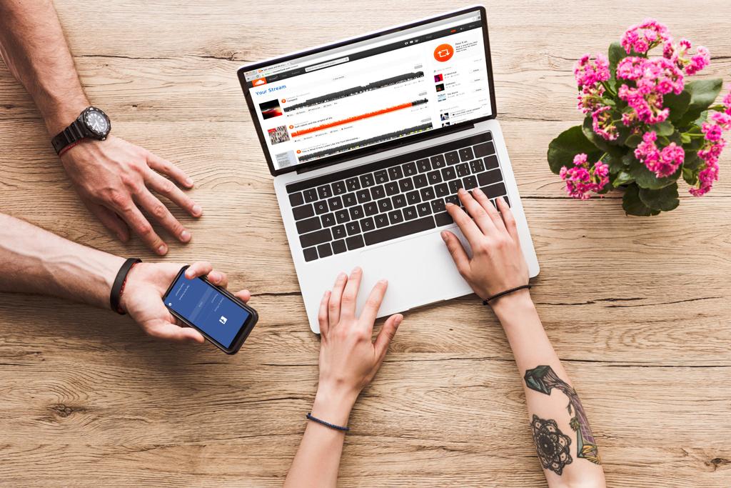 cropped shot of man with smartphone with facebook logo in hand and woman at tabletop with laptop with soundcloud website and kalanchoe flower - Photo, Image