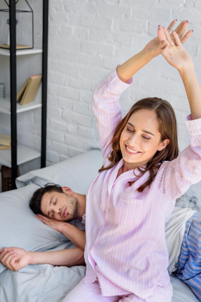Szczęśliwa młoda kobieta w piżamie, rozciągający się na łóżku, podczas gdy chłopak spanie za - Zdjęcie, obraz