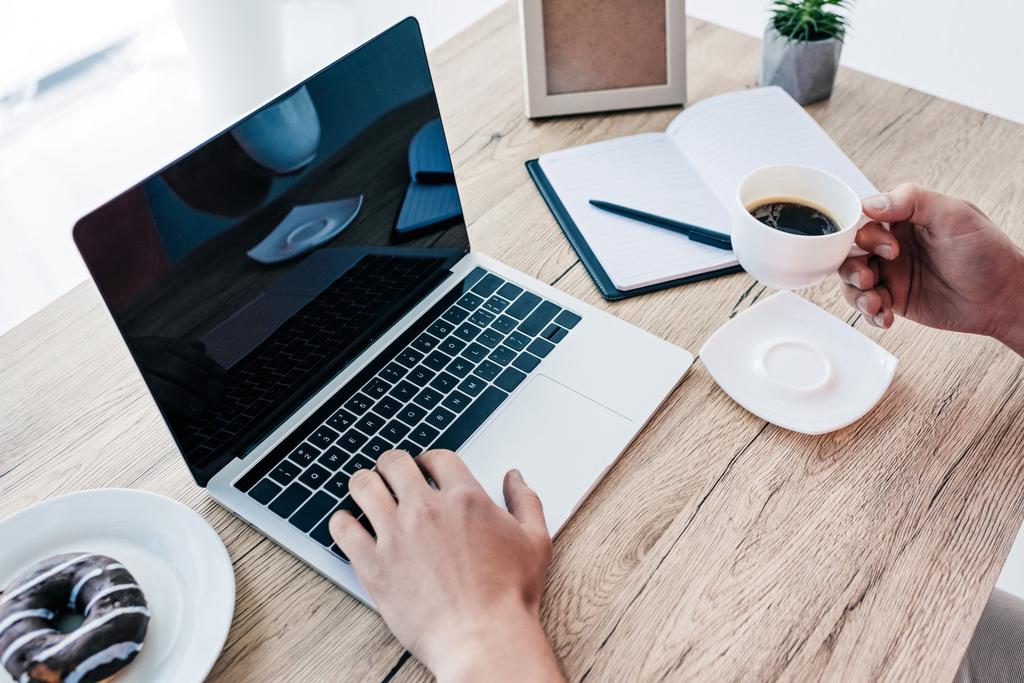 обрезанное изображение человека с чашкой кофе и пончиком за столом с ноутбуком, учебником, горшечным растением и фоторамкой
 - Фото, изображение