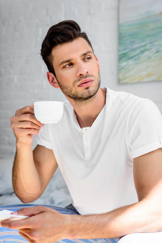 joven guapo en pijama sosteniendo una taza de café y mirando a casa
 - Foto, Imagen