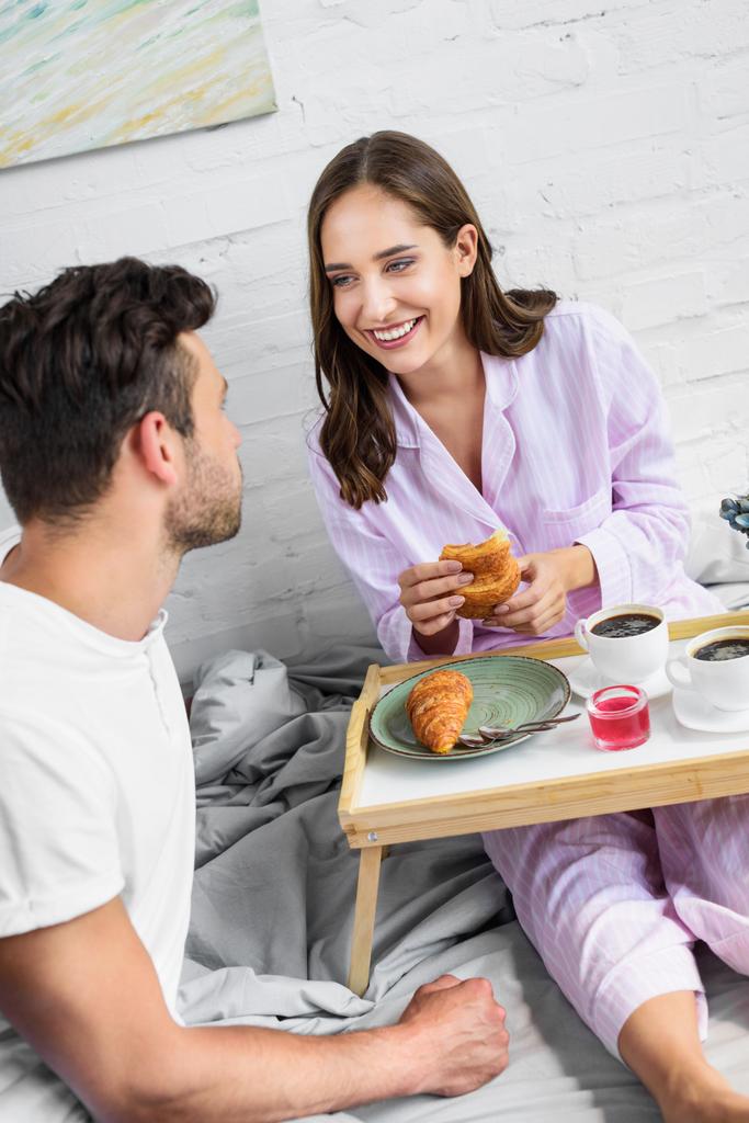 young laughing couple in pajamas having breakfast with coffee and croissants in bed together - Photo, Image