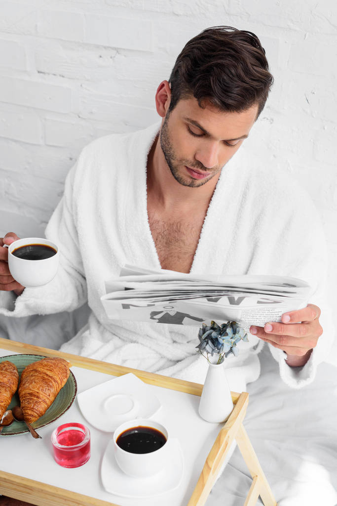 Молодой красивый мужчина в халате читает газету за завтраком с кофе и круассанами
 - Фото, изображение