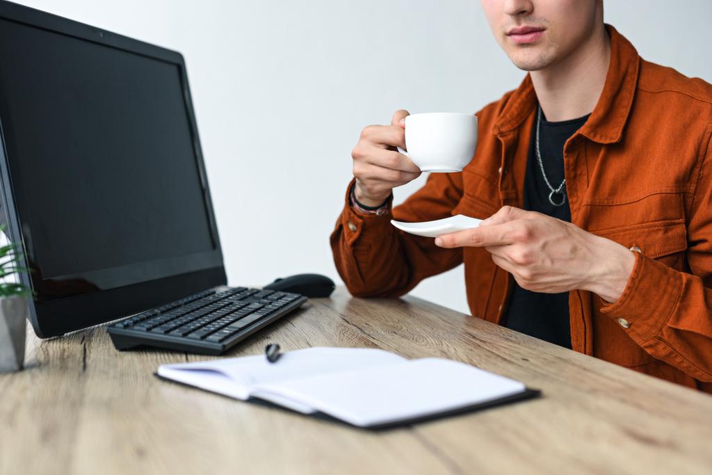 vue partielle de l'homme tenant tasse à café à table avec manuel, ordinateur, clavier d'ordinateur et souris d'ordinateur
 - Photo, image