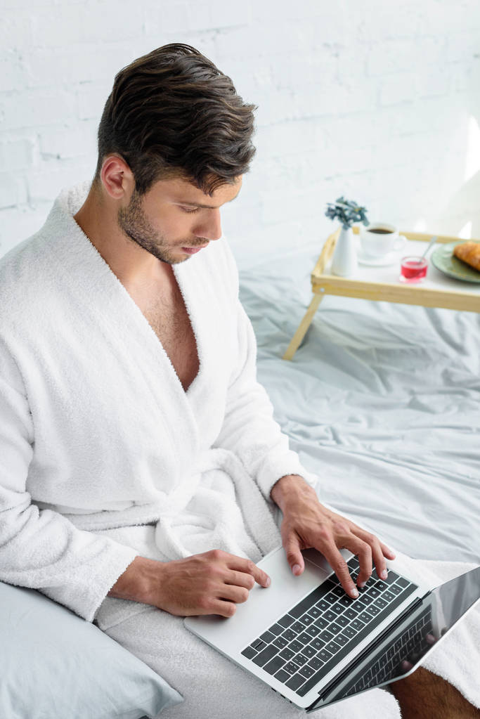 młody człowiek w szlafrok, siedząc na łóżku i komputera przenośnego - Zdjęcie, obraz