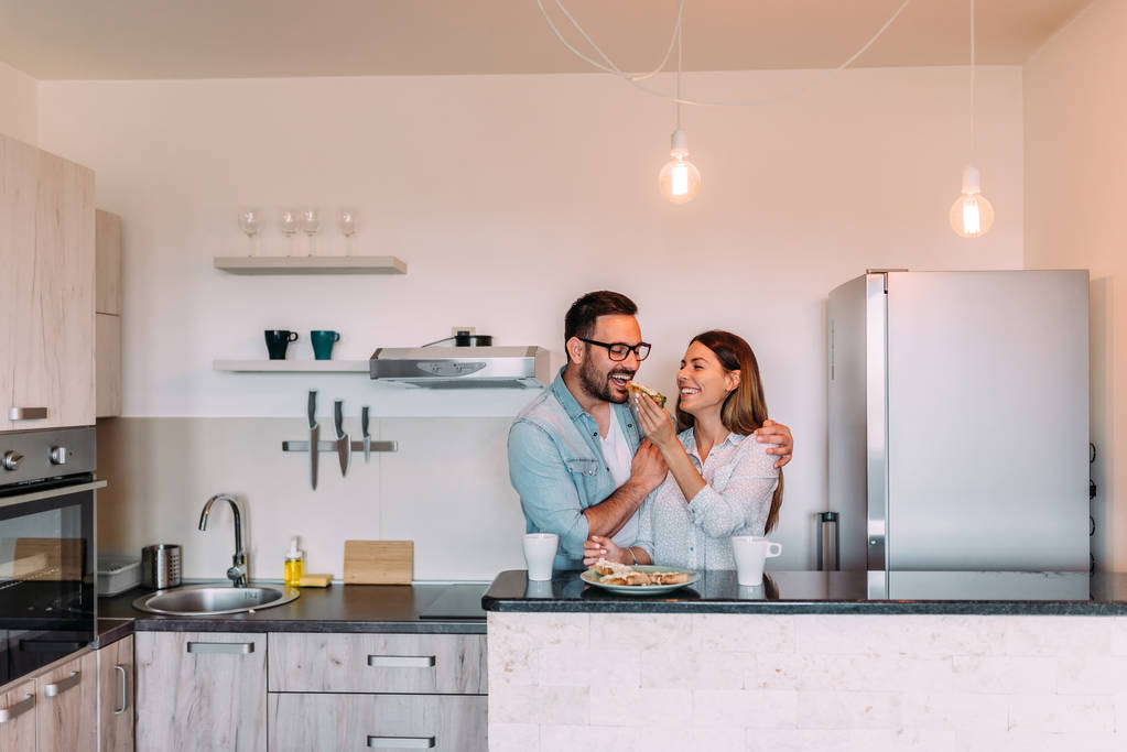 Ζευγάρι στέκεται στην κουζίνα του μοντέρνο διαμέρισμα και έχοντας πρωινό μαζί. - Φωτογραφία, εικόνα