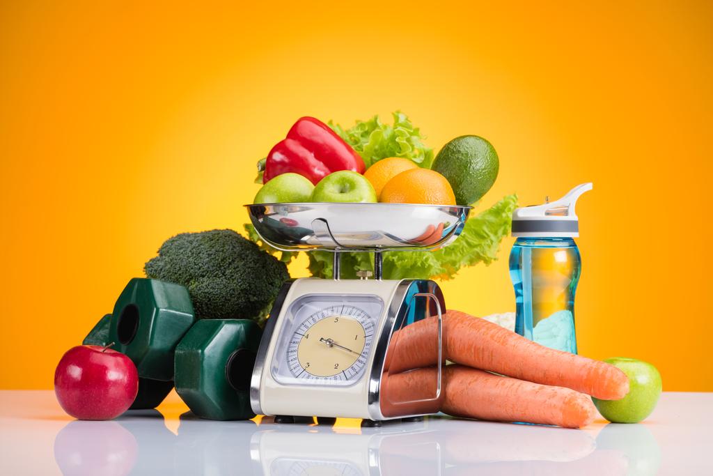 Nahaufnahme von frischem Obst und Gemüse auf der Waage, Hanteln und Wasserflasche auf gelb  - Foto, Bild