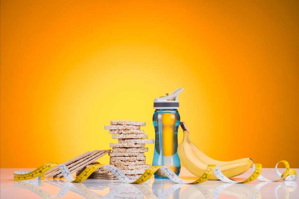 vista close-up de garrafa de esportes com água, biscoitos de dieta, fita métrica e banana no amarelo
 - Foto, Imagem