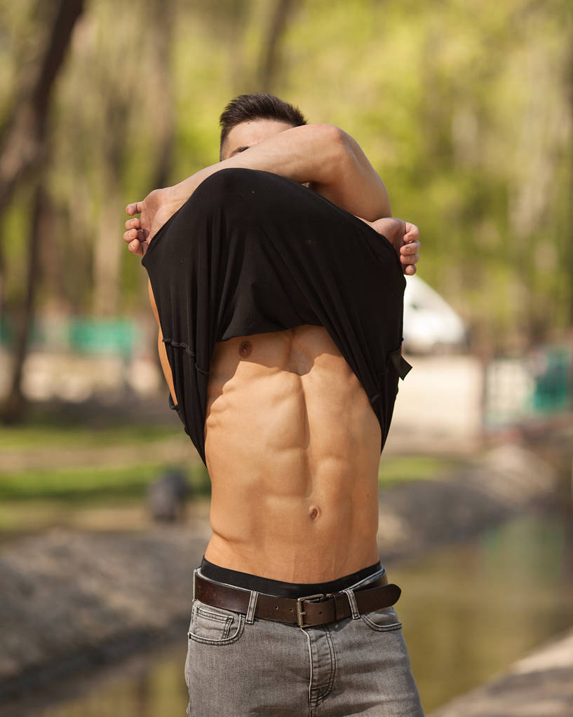 Velmi sexy mladý muž zobrazeno břišních svalů s sundat tričko na rozostřeného pozadí. Hezký, brutální macho v šortkách, při pohledu na fotoaparát. - Fotografie, Obrázek