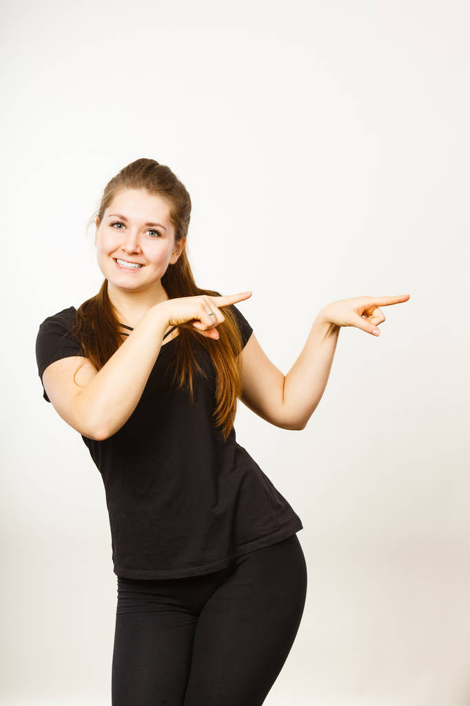 Ευτυχισμένος θετική γυναίκα που φοράει μαύρη στολή δείχνοντας κάτι με τα δάχτυλα. Studio που γυρίστηκε σε ανοιχτόχρωμο φόντο. - Φωτογραφία, εικόνα