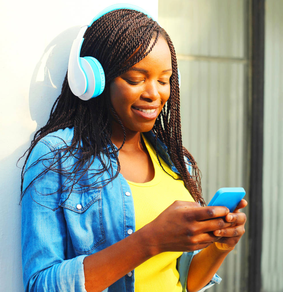ヘッドフォンと美しいアフリカ女性が市内でスマート フォンを使用して音楽を聴いてください。 - 写真・画像