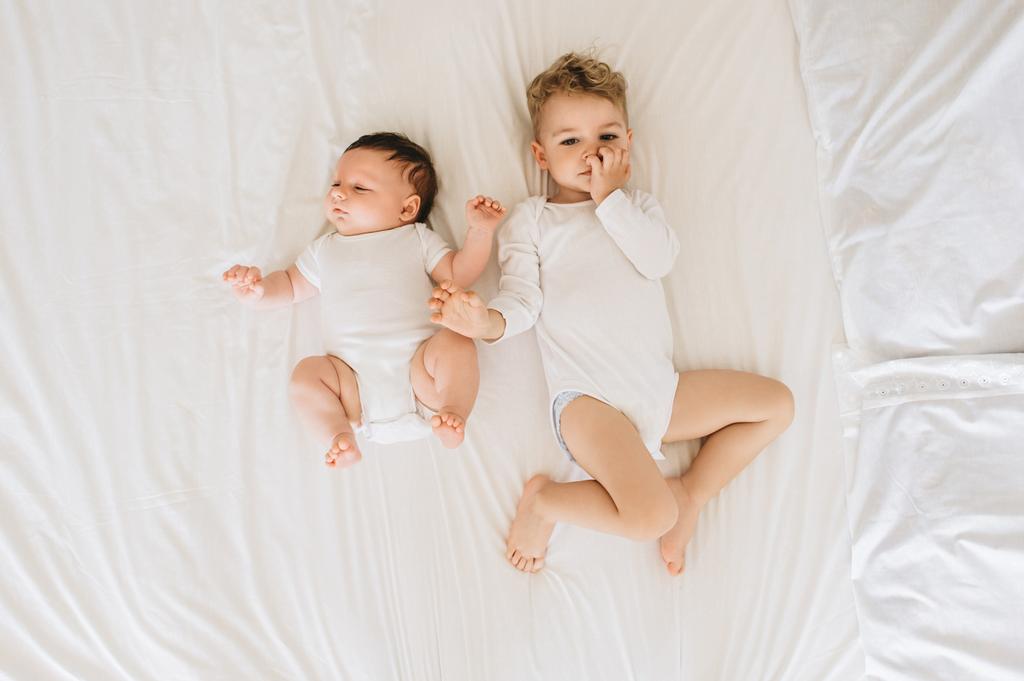 vista aerea di simpatici fratellini in body bianchi sdraiati sul letto insieme a casa
 - Foto, immagini