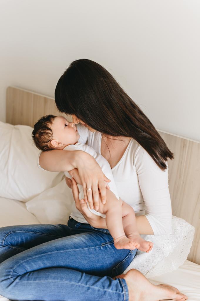 προσωπογραφία της μητέρας, κρατώντας λίγο βρέφος μωρό στα χέρια ενώ κάθεται στο κρεβάτι στο σπίτι - Φωτογραφία, εικόνα
