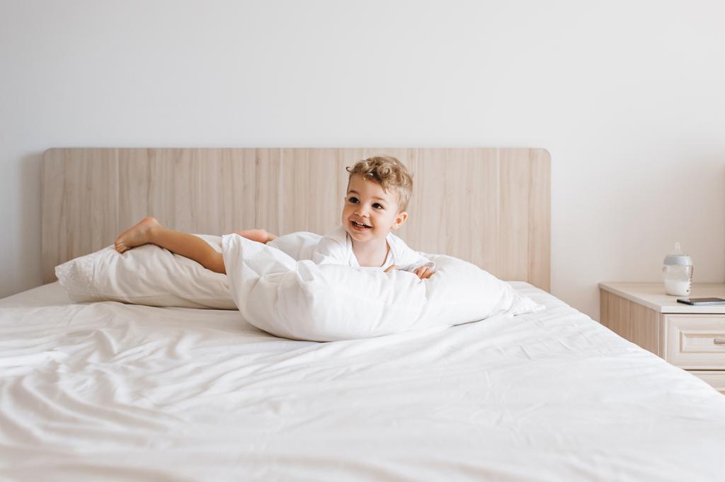 niño sonriente en traje de cuerpo blanco acostado en almohadas en la cama en casa
 - Foto, imagen