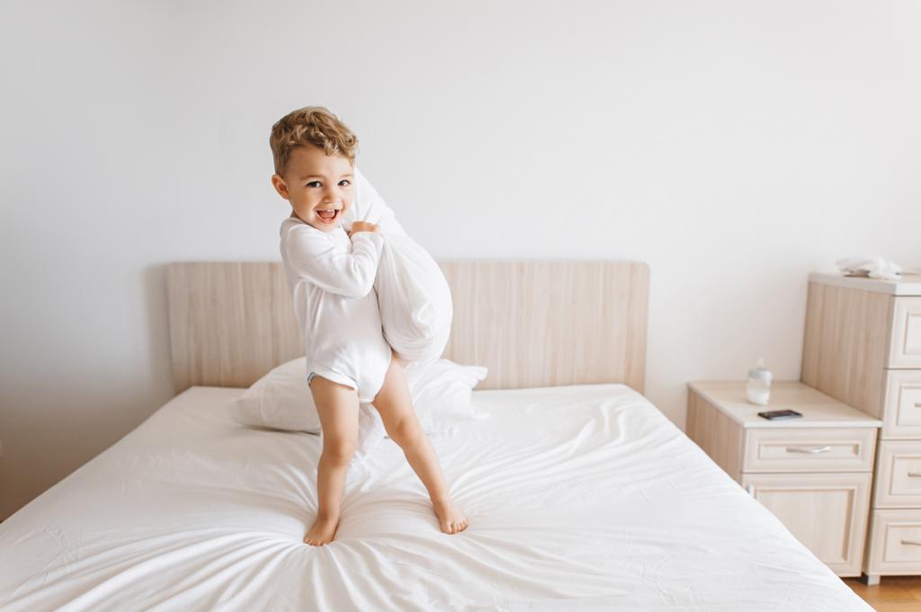 очаровательный мальчик в белом купальнике играет с подушкой на кровати дома
 - Фото, изображение