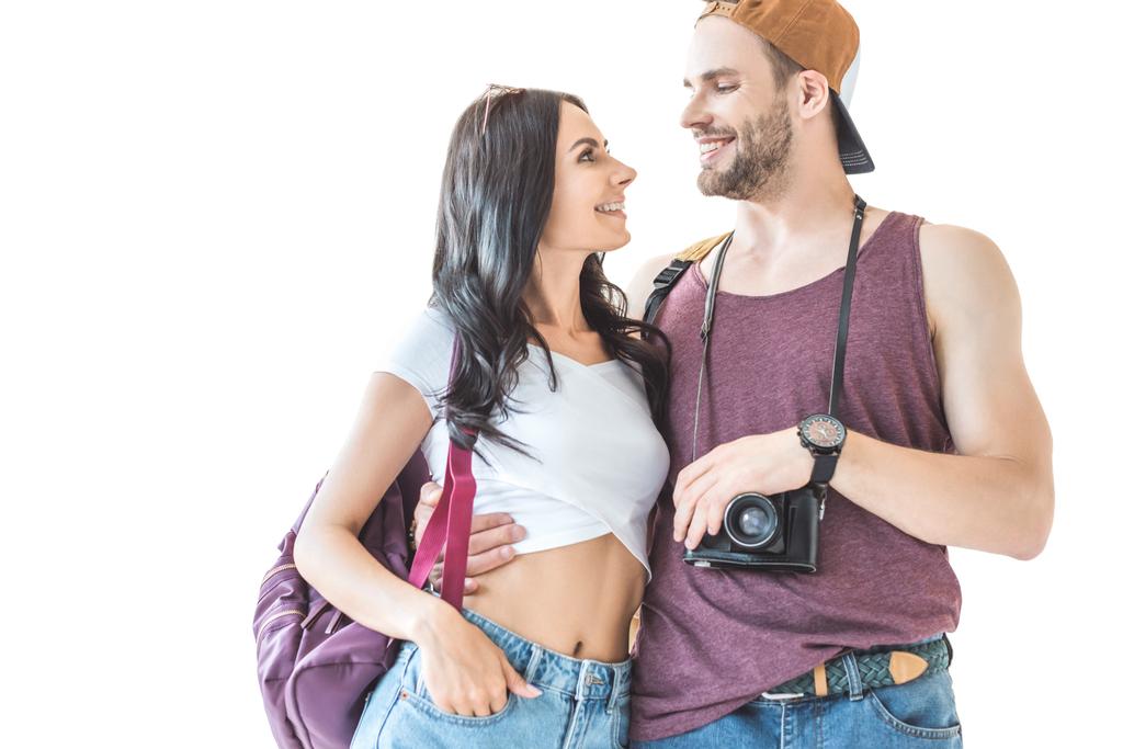 Ein paar lächelnde Touristen mit Kamera und Rucksack, die sich umarmen und einander anschauen, isoliert auf weiß - Foto, Bild