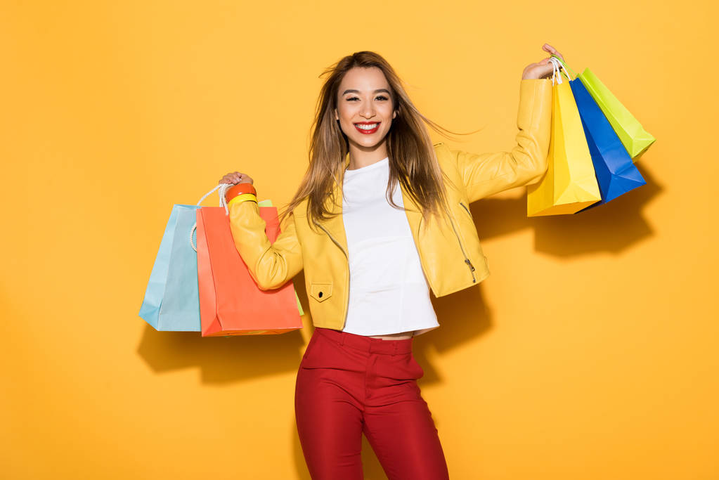 glimlachend stijlvolle Aziatische vrouw met boodschappentassen op gele achtergrond  - Foto, afbeelding
