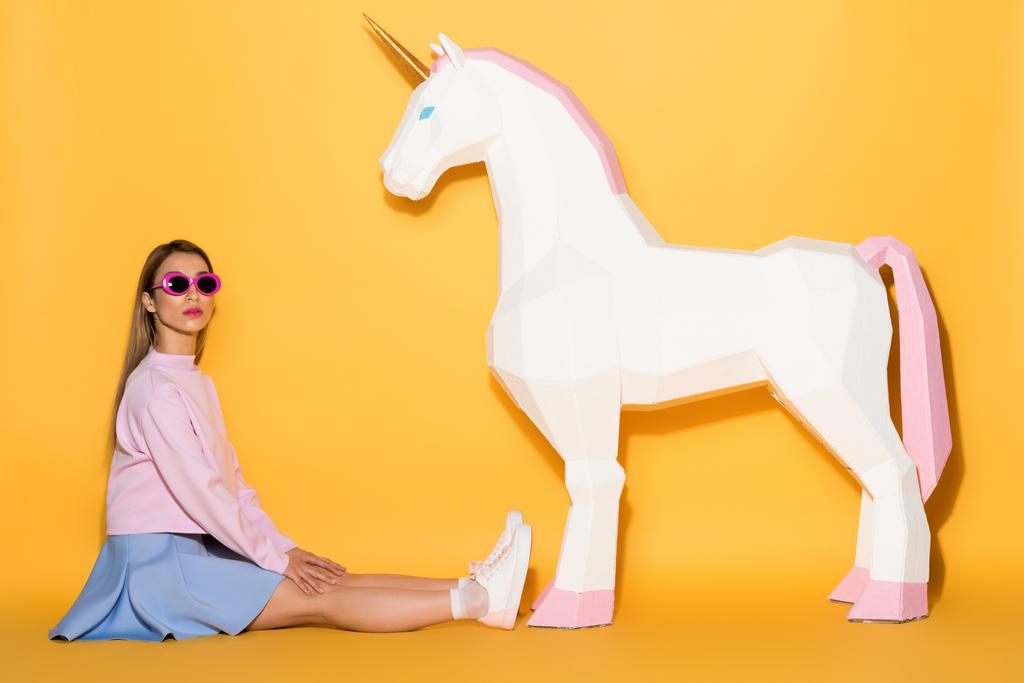 asian female model in sunglasses sitting on floor and decorative unicorn on yellow background  - Photo, Image