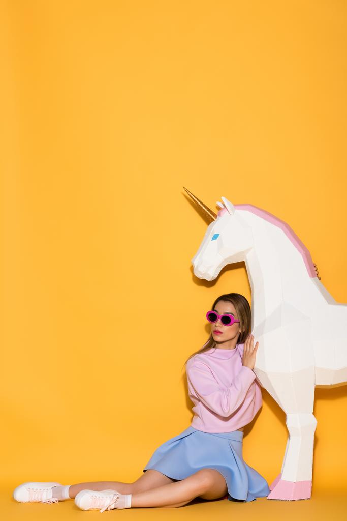 asian female model in sunglasses sitting on floor and decorative unicorn on yellow background - Photo, Image